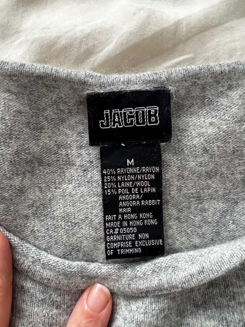 K&ouml;nnte beinhalten: Nahaufnahme eines grauen Pullovers mit einem schwarzen Etikett. Das Etikett zeigt "JACOB" in wei&szlig;en Blockbuchstaben. Darunter die Gr&ouml;&szlig;e "M" und eine Liste der Stoffe, darunter Viskose, Nylon, Wolle und Angora-Kaninchenhaar. Hergestellt in Hongkong.
