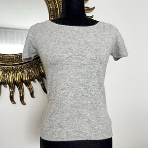 K&ouml;nnte beinhalten: Hellgraues Kurzarm-Shirt mit rundem Ausschnitt, pr&auml;sentiert auf einer schwarzen Schaufensterpuppe. Im Hintergrund ist ein goldgerahmter Spiegel zu sehen. Das Shirt ist aus weichem Material.
