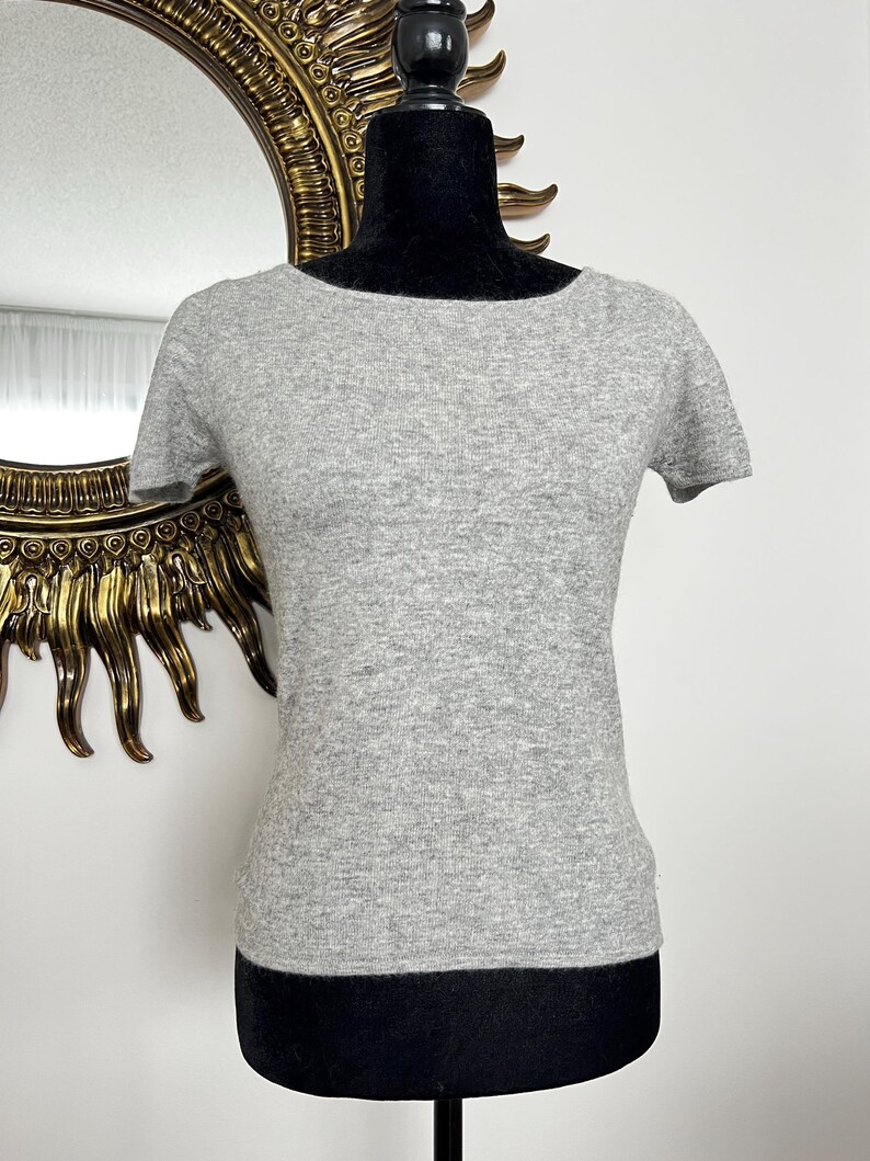 K&ouml;nnte beinhalten: Hellgraues Kurzarm-Shirt mit rundem Ausschnitt, pr&auml;sentiert auf einer schwarzen Schaufensterpuppe. Im Hintergrund ist ein goldgerahmter Spiegel zu sehen. Das Shirt ist aus weichem Material.