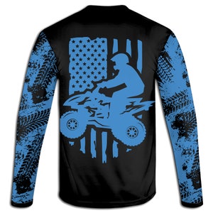 May include: Black long-sleeve shirt with a blue silhouette of a person riding an ATV in front of an American flag design. The sleeves have a blue and black tire tread pattern.