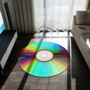 May include: A round rug designed to resemble a CD. The rug features a vibrant rainbow color scheme with a silver center. It is placed on a wooden floor in a living room setting, near a window and a sofa.