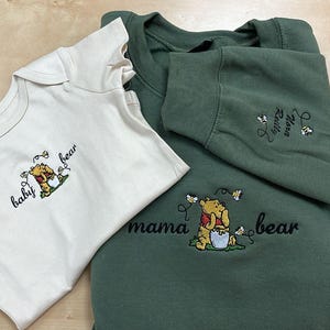 May include: An off-white baby onesie and a green sweatshirt with embroidered Winnie the Pooh designs. The onesie says "baby bear" and the sweatshirt says "mama bear". Both have a cartoon bear with a honey pot.