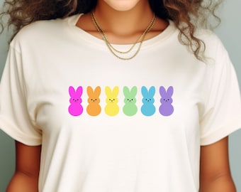 Könnte beinhalten: Ein weißes T-Shirt mit einer Reihe von sechs bunten, kaninchenförmigen Marshmallow-Peeps. Die Peeps sind in einem Regenbogenmuster angeordnet, beginnend mit rosa, orange, gelb, grün, blau und lila.