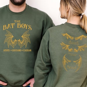 Puede incluir: Dos sudaderas de color verde oliva con un estampado dorado de alas de murci&eacute;lago y el texto "The Bat Boys" y los nombres "Azriel, Rhysand, Cassian".