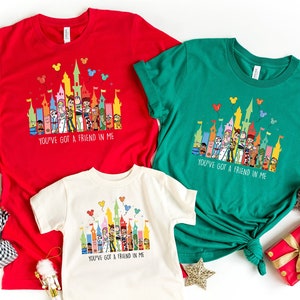 Op de afbeelding: Drie T-shirts met een kleurrijke illustratie van een kasteel met personages uit de film Toy Story. De shirts zijn rood, groen en wit. De tekst "You've got a friend in me" staat op elk shirt gedrukt.