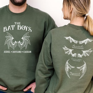 Puede incluir: Una sudadera verde con un estampado blanco de alas de murci&eacute;lago y el texto "The Bat Boys" y "Azriel + Rhysand + Cassian". La parte posterior de la sudadera tiene tres conjuntos de alas de murci&eacute;lago con los nombres "Azriel", "Cassian" y "Rhysand" escritos debajo.