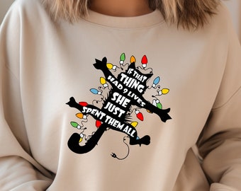May include: A beige sweatshirt with a black cat silhouette decorated with colorful Christmas lights. The text on the sweatshirt reads, "If that thing had 9 lives she just spent them all."