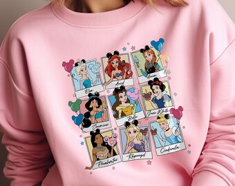 May include: Pink sweatshirt featuring a graphic print of Disney princesses in Polaroid frames with balloons. The princesses include Elsa, Ariel, Anna, Jasmine, Belle, Snow White, Pocahontas, Rapunzel, and Cinderella.