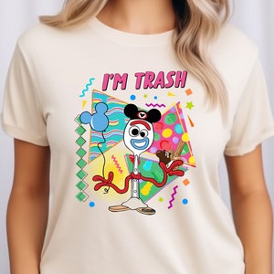 May include: A white t-shirt featuring a cartoon image of a toy spoon wearing a Mickey Mouse hat and holding a balloon. The text "I'M TRASH" is printed above the image.