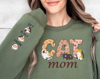 Puede incluir: Sudadera verde con el texto "CAT MOM" en letras marrones. Las letras están decoradas con un patrón de gatos. La manga izquierda tiene el texto "Mimi, Kona, Lily" con huellas de patas.