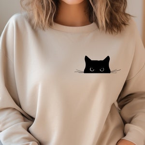 May include: A beige sweatshirt with a black cat peeking out from behind the fabric. The cat has large eyes and a small nose.