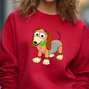 May include: Red sweatshirt with a cartoon image of Slinky Dog from the Toy Story franchise. The dog is brown and gray with a spring body and a green collar.
