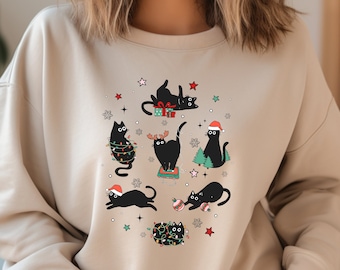 May include: Beige sweatshirt with a Christmas-themed design of black cats. The cats are adorned with festive decorations like fairy lights, Santa hats, and gifts. The design includes stars and snowflakes, creating a playful and cosy holiday look.