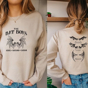 Puede incluir: Una sudadera beige con un estampado gr&aacute;fico negro en la parte delantera que dice "THE BAT BOYS AZRIEL + RHYSAND + CASSIAN". La parte posterior de la sudadera tiene tres gr&aacute;ficos de alas de murci&eacute;lago negras con los nombres "Azriel", "Cassian" y "Rhysand" escritos debajo de cada uno.