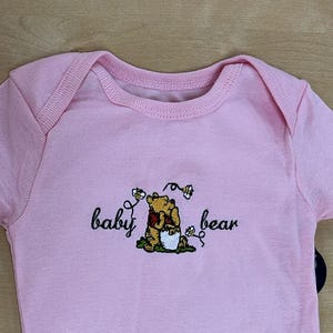 May include: A light pink baby onesie with short sleeves and a snap closure. The onesie features an embroidered design of a bear with the words "baby bear". The Gerber brand label is visible.