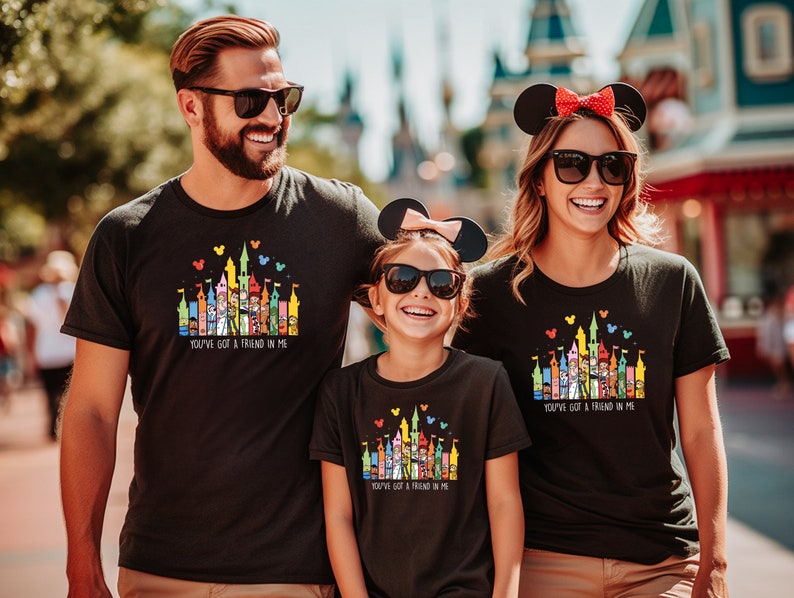 Op de afbeelding: Een gezin van drie personen draagt zwarte T-shirts met een kleurrijke illustratie van het Disney-kasteel en de tekst "You've got a friend in me".