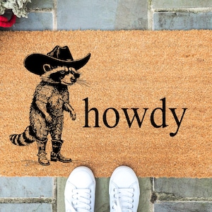 May include: A rectangular doormat with a brown coir surface. A raccoon wearing a cowboy hat and boots is printed on the mat. The word "howdy" is printed in black. White sneakers are at the bottom of the image.