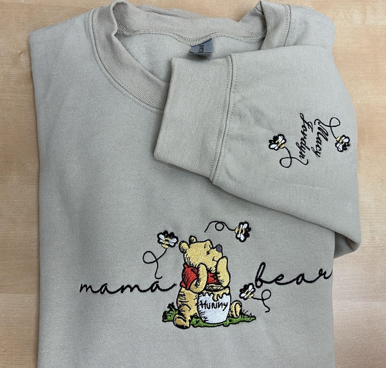 May include: A beige sweatshirt with embroidered Winnie the Pooh graphics. The design includes the words "mama bear" and "Hunny", along with the names "Macy" and "Jordyn" on the sleeve.
