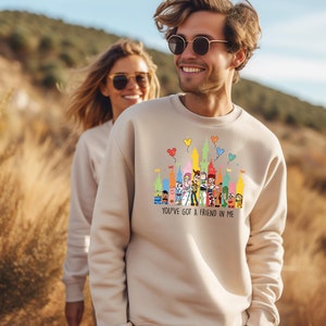 Op de afbeelding: Een beige sweatshirt met een kleurrijke afbeelding van Disney-figuren voor een kasteel. De tekst "You've got a friend in me" staat onder de afbeelding.