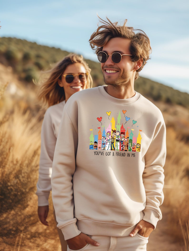 Op de afbeelding: Een beige sweatshirt met een kleurrijke afbeelding van Disney-figuren voor een kasteel. De tekst "You've got a friend in me" staat onder de afbeelding.