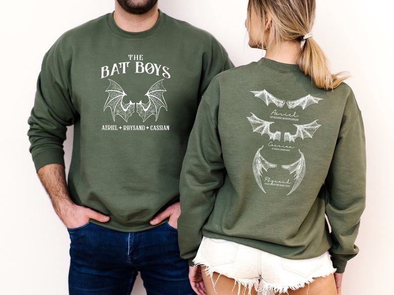 Puede incluir: Una sudadera verde con un estampado blanco de alas de murci&eacute;lago y el texto "The Bat Boys" y "Azriel + Rhysand + Cassian". La parte posterior de la sudadera tiene tres conjuntos de alas de murci&eacute;lago con los nombres "Azriel", "Cassian" y "Rhysand" escritos debajo.