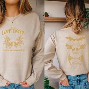 Puede incluir: Una sudadera beige con un estampado gr&aacute;fico dorado de alas de murci&eacute;lago y el texto "THE BAT BOYS" y "AXRIEL + RHYSAND + CASSIAN". La parte posterior de la sudadera tiene tres dise&ntilde;os de alas de murci&eacute;lago doradas con los nombres "AXRIEL", "CASSIAN" y "RHYSAND" escritos debajo de cada uno.