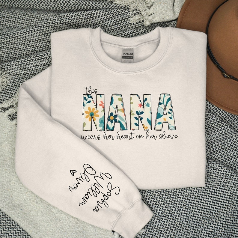 Op de afbeelding: Witte trui met de tekst "This Nana wears her heart on her sleeve" in zwarte letters. Het woord "Nana" staat in grote, kleurrijke letters met een bloemenpatroon.