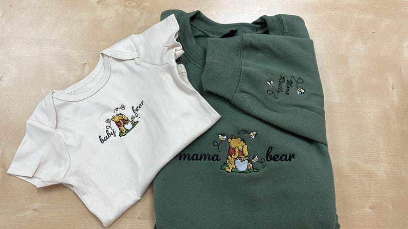 May include: An off-white baby onesie and a green sweatshirt with embroidered Winnie the Pooh designs. The onesie says "baby bear" and the sweatshirt says "mama bear". Both have a cartoon bear with a honey pot.