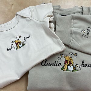 May include: A cream-colored baby onesie and a beige sweatshirt with embroidered Winnie the Pooh designs. The onesie says "baby bear" and the sweatshirt says "auntie bear". The sweatshirt also has the word "love" embroidered.