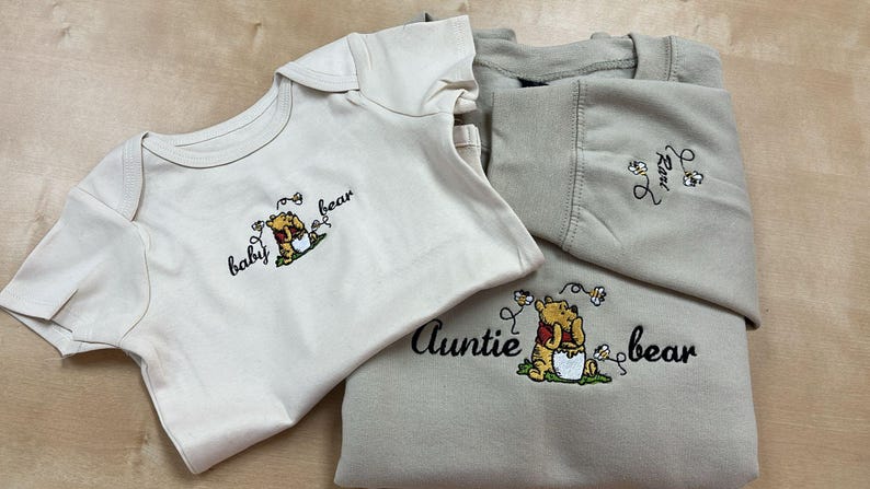 May include: A cream-colored baby onesie and a beige sweatshirt with embroidered Winnie the Pooh designs. The onesie says "baby bear" and the sweatshirt says "auntie bear". The sweatshirt also has the word "love" embroidered.