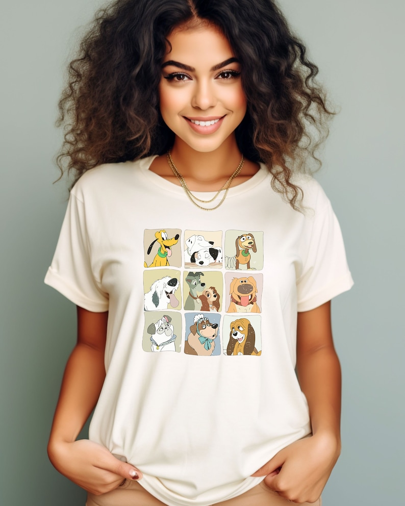 Pu&ograve; includere: T-shirt color crema con una griglia di nove illustrazioni di cani dei cartoni animati. I cani sono in singoli quadrati con un bordo marrone chiaro. La maglietta ha un girocollo classico e maniche corte.