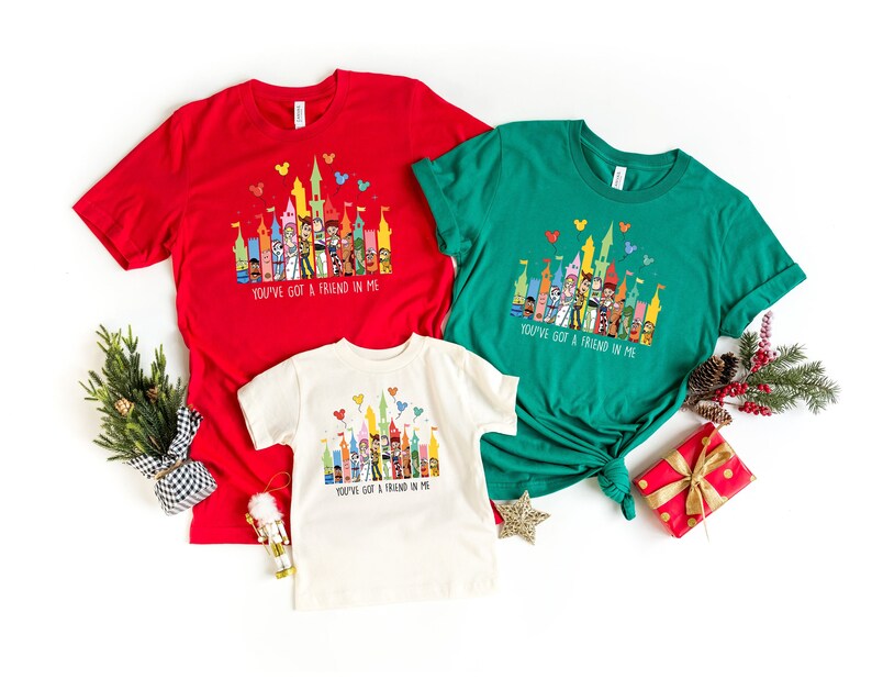 Op de afbeelding: Drie T-shirts met een kleurrijke illustratie van een kasteel met personages uit de film Toy Story. De shirts zijn rood, groen en wit. De tekst "You've got a friend in me" staat op elk shirt gedrukt.