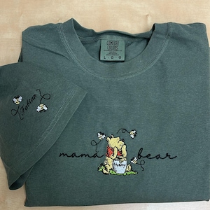 May include: A dark green t-shirt with the words "mama bear" and a Winnie the Pooh design embroidered on the front. The sleeve has the name "Fabian" and two bees embroidered on it. The shirt has a Comfort Colors tag.