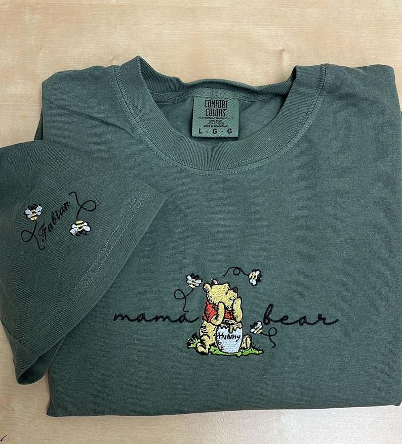 May include: A dark green t-shirt with the words "mama bear" and a Winnie the Pooh design embroidered on the front. The sleeve has the name "Fabian" and two bees embroidered on it. The shirt has a Comfort Colors tag.