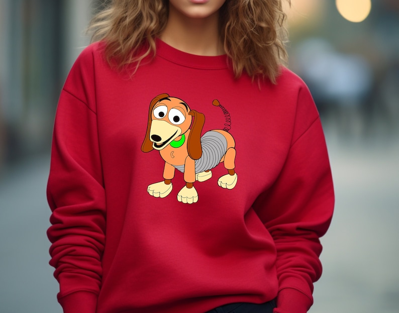 May include: Red sweatshirt with a cartoon image of Slinky Dog from the Toy Story franchise. The dog is brown and gray with a spring body and a green collar.