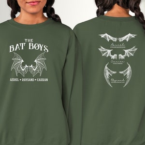 Puede incluir: Una sudadera verde con un estampado blanco de alas de murci&eacute;lago y el texto "THE BAT BOYS AZRIEL + RHYSAND + CASSIAN" en la parte delantera. La parte posterior de la sudadera tiene un estampado blanco de tres conjuntos de alas de murci&eacute;lago con los nombres "Azriel", "Cassian" y "Rhysand" escritos debajo de cada conjunto.