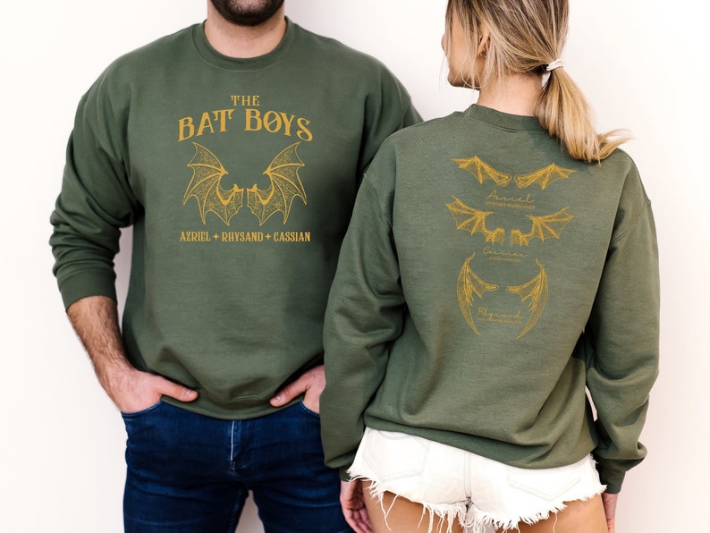 Puede incluir: Dos sudaderas de color verde oliva con un estampado dorado de alas de murci&eacute;lago y el texto "The Bat Boys" y los nombres "Azriel, Rhysand, Cassian".