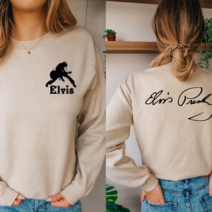 May include: A beige sweatshirt with a black silhouette of Elvis Presley playing a guitar on the front and his signature on the back. The text "Elvis" is printed below the silhouette.