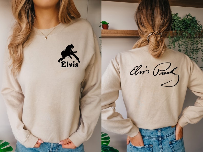 May include: A beige sweatshirt with a black silhouette of Elvis Presley playing a guitar on the front and his signature on the back. The text "Elvis" is printed below the silhouette.