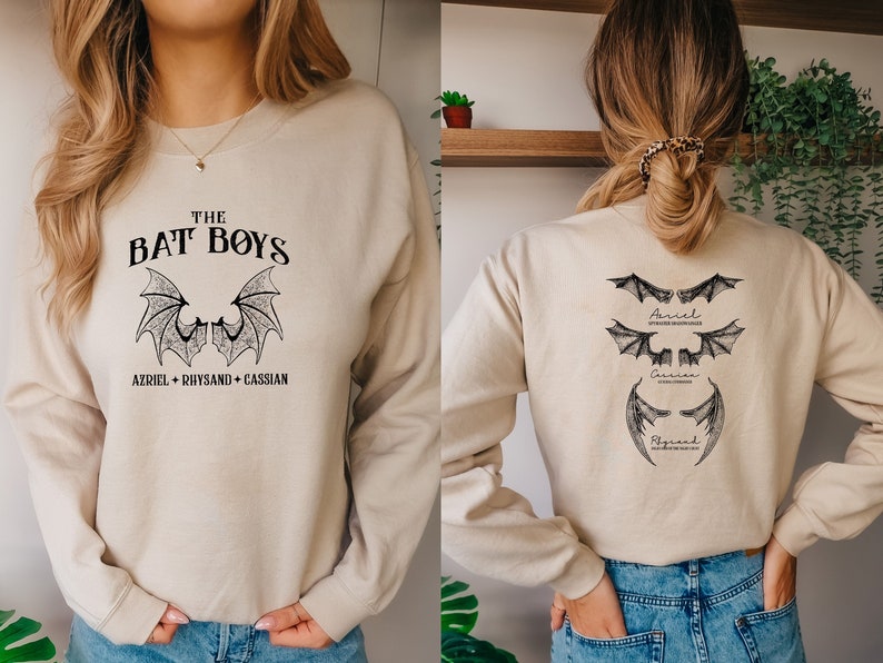 Puede incluir: Una sudadera beige con un estampado gr&aacute;fico negro en la parte delantera que dice "THE BAT BOYS AZRIEL + RHYSAND + CASSIAN". La parte posterior de la sudadera tiene tres gr&aacute;ficos de alas de murci&eacute;lago negras con los nombres "Azriel", "Cassian" y "Rhysand" escritos debajo de cada uno.