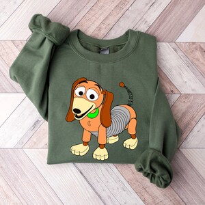 May include: Green sweatshirt with a cartoon image of Slinky Dog from the Toy Story franchise. Slinky Dog is orange and brown with a spring body and a green collar.