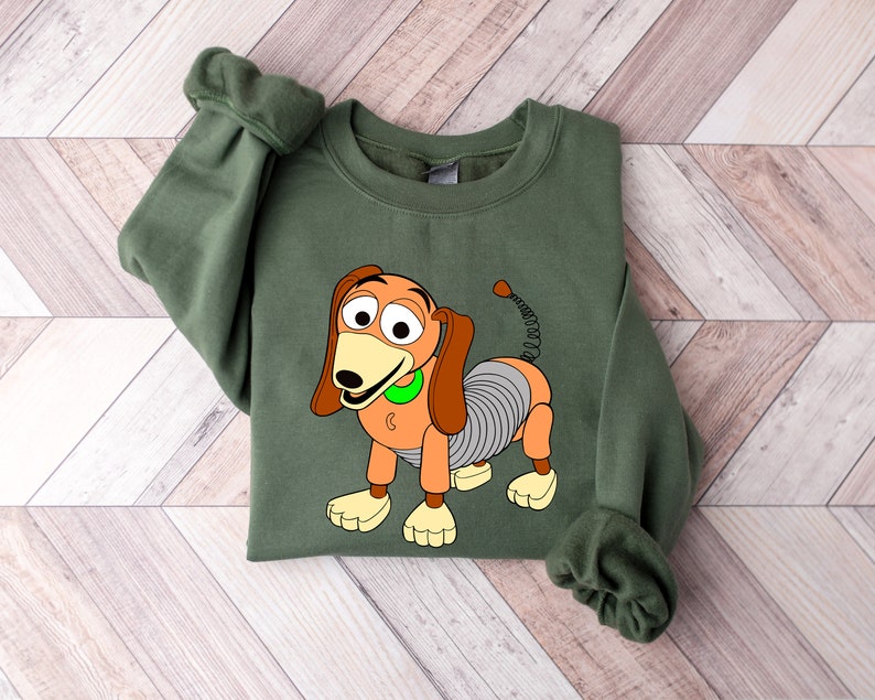 May include: Green sweatshirt with a cartoon image of Slinky Dog from the Toy Story franchise. Slinky Dog is orange and brown with a spring body and a green collar.