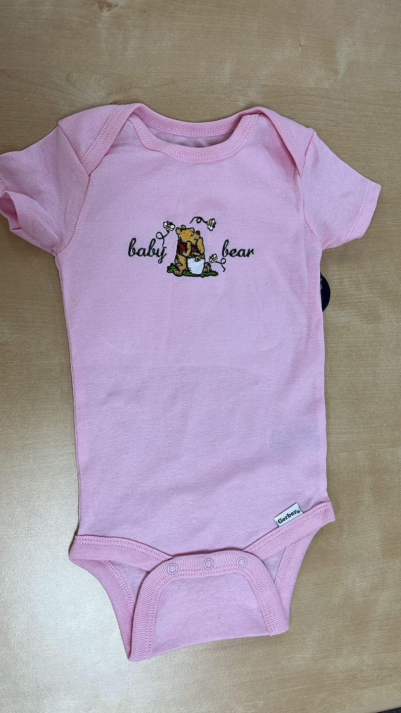 May include: A light pink baby onesie with short sleeves and a snap closure. The onesie features an embroidered design of a bear with the words "baby bear". The Gerber brand label is visible.