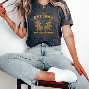 Puede incluir: Una camiseta gris con un gr&aacute;fico dorado de dos alas de murci&eacute;lago y el texto "THE BAT BOYS AZRIEL + RHYSAND + CASSIAN".