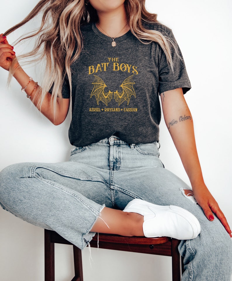 Puede incluir: Una camiseta gris con un gr&aacute;fico dorado de dos alas de murci&eacute;lago y el texto "THE BAT BOYS AZRIEL + RHYSAND + CASSIAN".