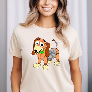 May include: A white t-shirt with a cartoon image of a brown and orange toy dog with a spring tail. The dog has a green collar and is wearing a smile.