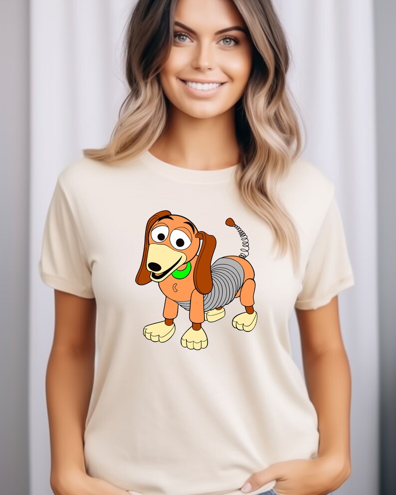 May include: A white t-shirt with a cartoon image of a brown and orange toy dog with a spring tail. The dog has a green collar and is wearing a smile.