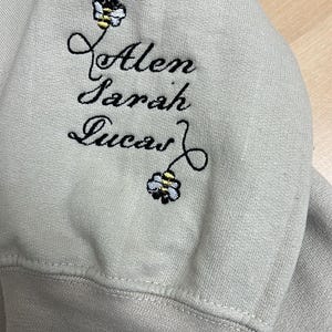 May include: A light gray sweatshirt sleeve with embroidered names: Alen, Sarah, and Lucas. Each name is accompanied by a small bee design. The embroidery is in black thread.