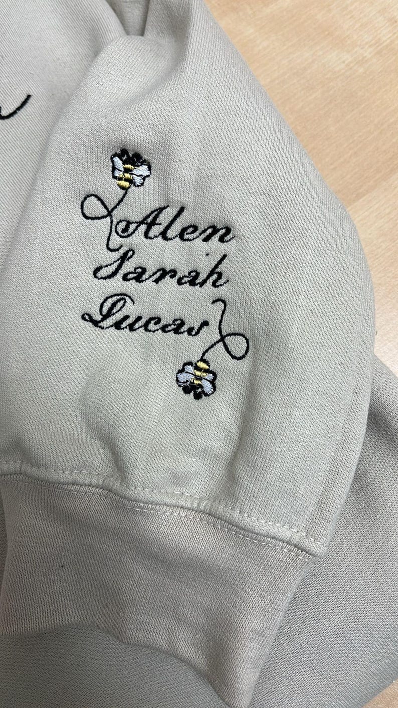 May include: A light gray sweatshirt sleeve with embroidered names: Alen, Sarah, and Lucas. Each name is accompanied by a small bee design. The embroidery is in black thread.