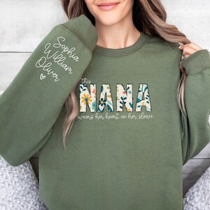 Op de afbeelding: Een groen sweatshirt met de tekst "This Nana wears her heart on her sleeve" en een bloemenontwerp. De mouw heeft de namen "Sophia, William, Oliver" erop geschreven.